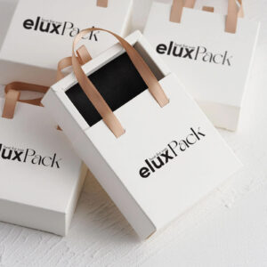 White cardboard gift boxes with ribbon handles, resembling elegant shopping bags. The boxes have a sleek design with a matte finish, and gold ribbon handles add a touch of luxury.